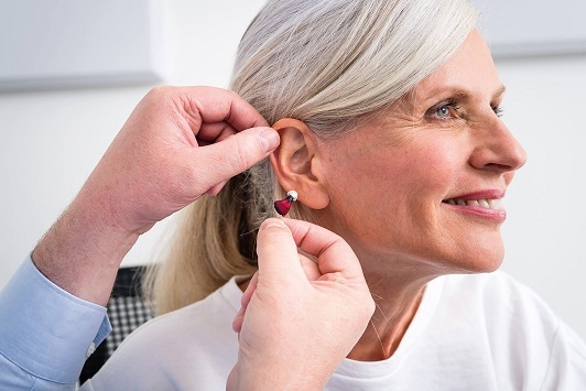 Eine ältere Frau bekommt ein modernes Hörgerät ins Ohr eingesetzt und lächelt zufrieden.